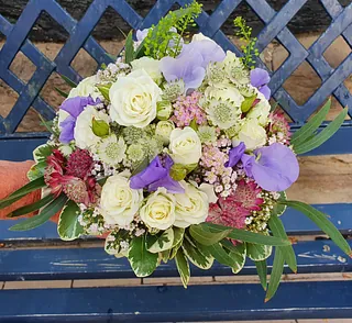 Bunter Blumenstrauß mit weißen Rosen, lila und rosa Blüten sowie grünem Laub auf einer blauen Bank vor einem Gitter