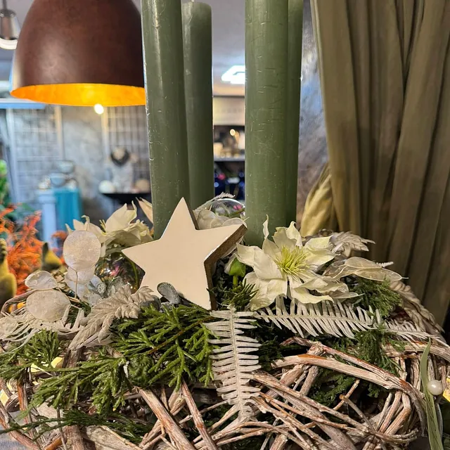 Adventskranz mit drei grünen Kerzen, einem weißen Stern, weißen Blüten und grünen Zweigen in einem Raum mit Vorhang und Lampe im Hintergrund