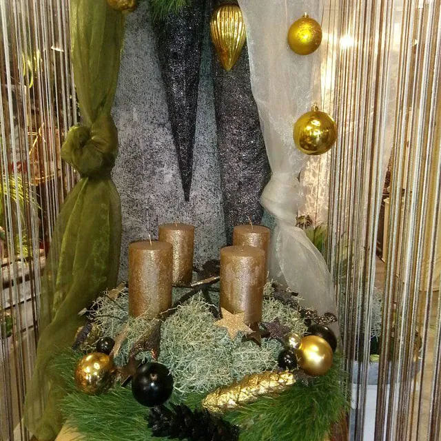 Adventskranz mit vier goldfarbenen Kerzen, goldenen und schwarzen Kugeln sowie goldfarbenen Dekorationen vor grünem und weißem Stoffhintergrund