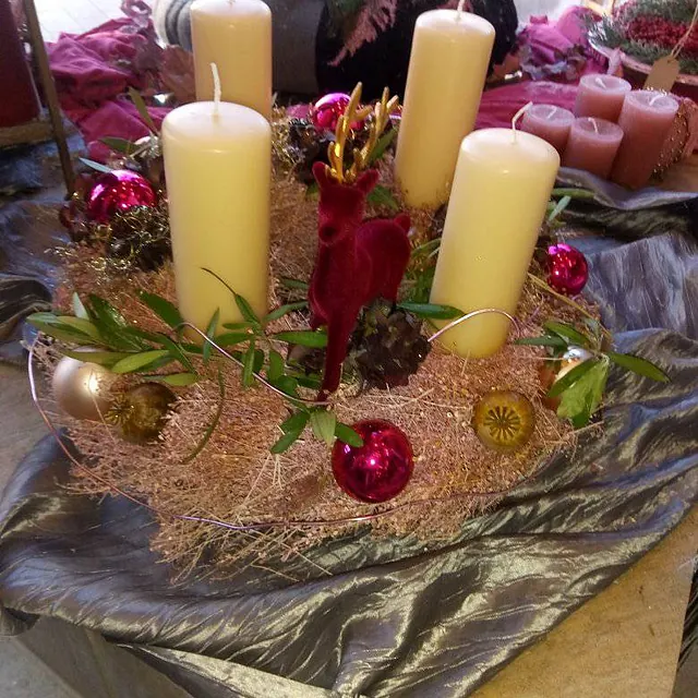Adventskranz mit vier großen weißen Kerzen, roten und goldenen Kugeln, grünen Zweigen und einem kleinen roten Rentier in der Mitte