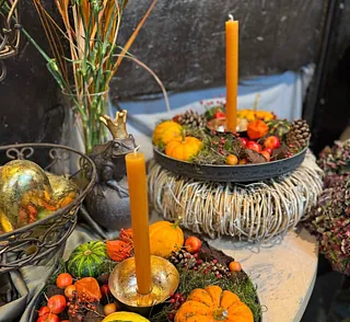 Zwei orangefarbene Kerzen in dekorativen Kränzen mit kleinen Kürbissen, Tannenzapfen und Beeren auf einem Tisch neben einer Metallfigur mit Krone und einem Drahtkorb mit goldfarbenen Kugeln