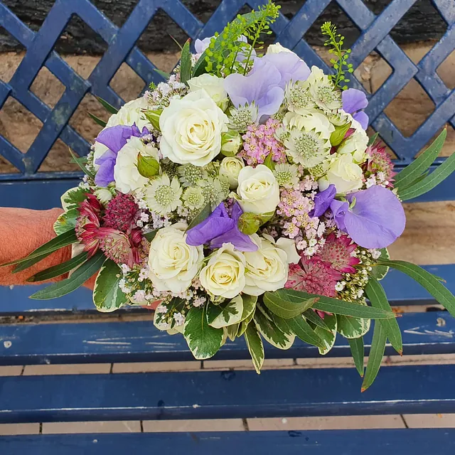 Bunter Blumenstrauß mit weißen Rosen, lila Blüten und grünem Laub auf einer blauen Holzbank