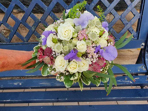 Blumenstrauß mit weißen Rosen, lila und rosa Blüten sowie grünem Laub vor blauem Metallgitter und Bank
