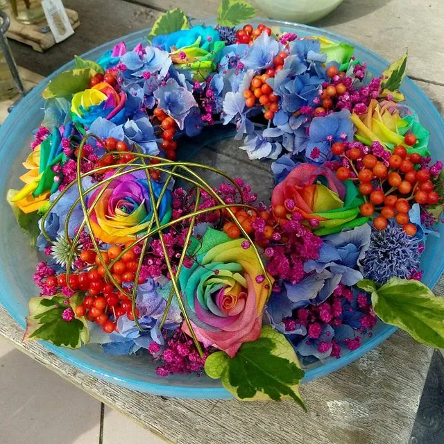 Bunter Blumenkranz mit regenbogenfarbenen Rosen, blauen Hortensien, roten Beeren und grünen Blättern auf einem blauen Teller